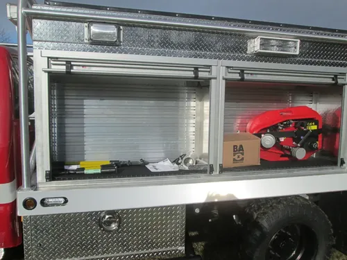 Exterior view of small fire truck showing cab, body compartments, and wheel/tire area.