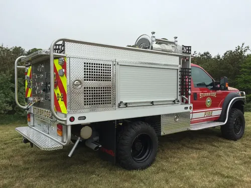 Exterior view of small fire truck showing cab, body compartments, and wheel/tire area.