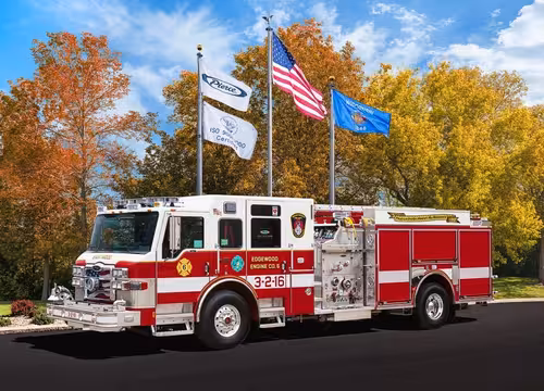 Front-left profile of a pumper beneath flagpoles showing cab face, pump panel section, and rear body.