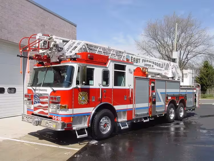Front-left exterior view of a ladder truck with stowed aerial, side compartments, and tandem rear axles.
