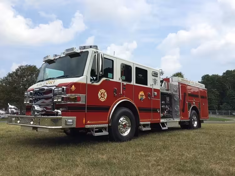 Front-left exterior view of a pumper showing the cab, bumper, pump panel area, and side compartments.
