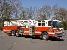 Wide front right view of the parked tower ladder with cab, stowed aerial, and rear body compartments.