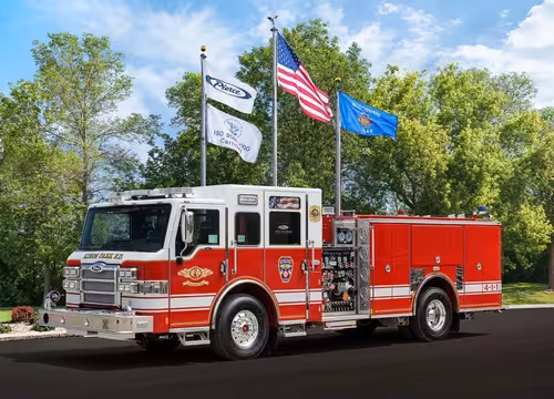 Front-left profile view with the pumper parked beneath flagpoles showing full side body and wheelbase.