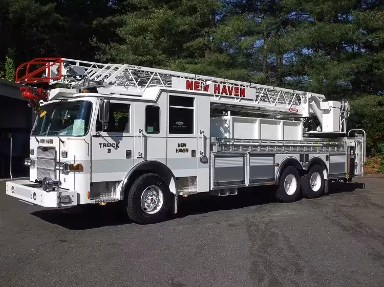 Front-left exterior view of a ladder truck with stowed aerial, side compartments, and tandem rear axles.