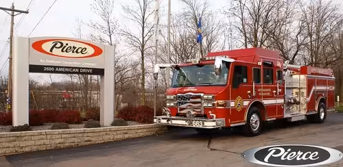 Front-left wide view of the pumper at the Pierce sign with side pump panel section visible.