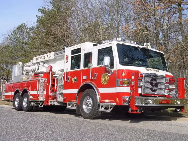 Front-right exterior view of a rear-mount aerial ladder with front outriggers, side compartments, and tandem rear axles.