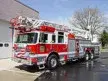 Wide front-left view of the ladder truck parked by the bay with stowed aerial visible.