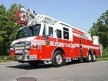 Wide front-right view of the aerial ladder parked roadside with front outrigger and tandem rear axles visible.