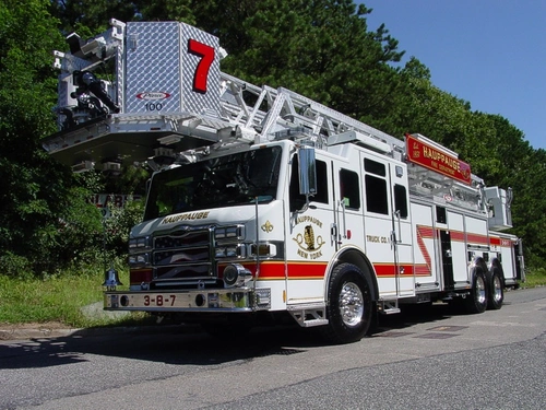 Front-left exterior view of a ladder truck with stowed aerial, side compartments, and tandem rear axles.