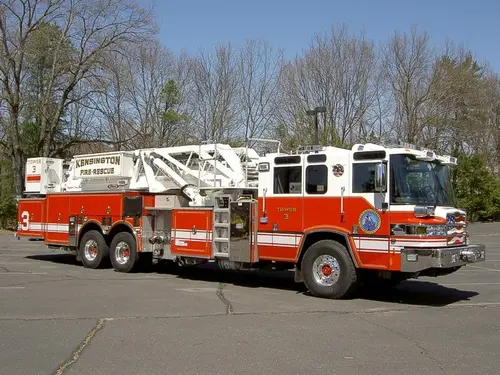 Front right exterior view of the rear-mount tower ladder with tandem rear axles, body compartments, and stowed aerial.