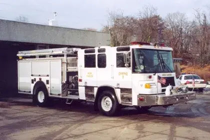 Front right exterior view of a white pumper with chrome bumper, cab roof lights, and side compartment body.