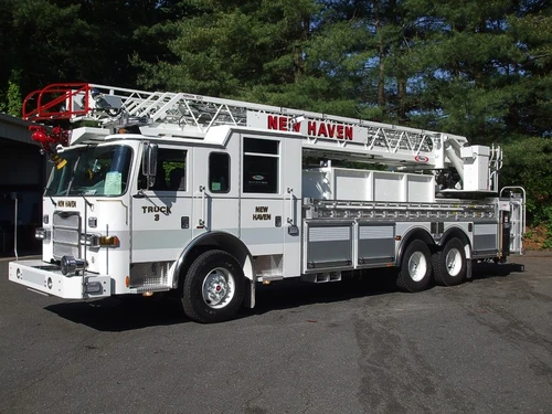 Front-left exterior view of a ladder truck with stowed aerial, side compartments, and tandem rear axles.