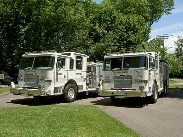 Front left view of two white pumpers parked side by side with chrome bumpers and roof warning lights.