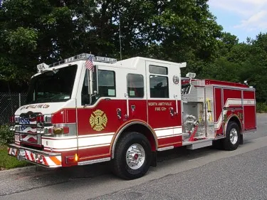 Front-left exterior view of a pumper showing the cab, bumper, pump panel area, and side compartments.