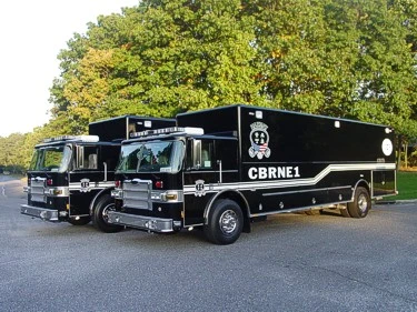 Front left view of two black hazmat support trucks parked side by side with large box bodies and lightbars.