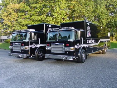 Front three-quarter view of both hazmat trucks showing chrome grilles, bumpers, and long command-style bodies.
