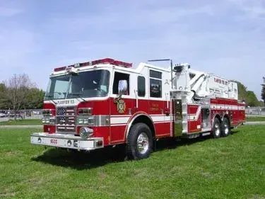 Front-left exterior view of tower ladder truck parked on grass with ladder stowed
