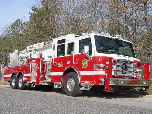 Front-right exterior view of a rear-mount aerial ladder with front outriggers, side compartments, and tandem rear axles.