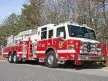 Wide front-right view of the aerial ladder parked roadside with the ladder bed and tandem rear wheels visible.